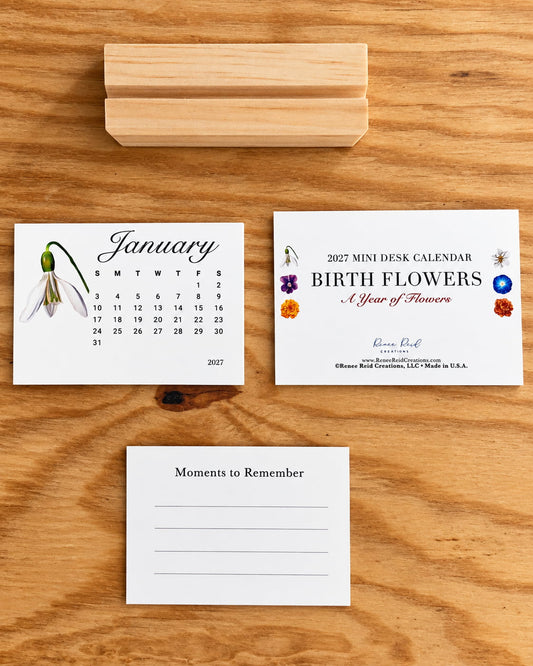 2027 mini desk calendar featuring hand-drawn birth flower illustrations by Renee Reid Creations, displayed with a January snowdrop calendar card, floral cover card, and a "Moment to Remember" note card on a light wood surface.
