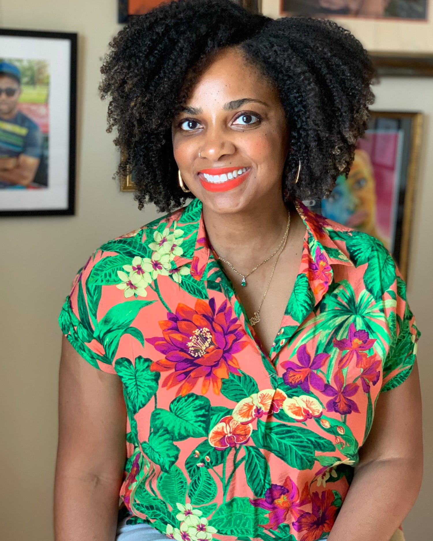 Picture of a smiling woman (Colored Pencil Artist Renee Reid of Renee Reid Creations) with curly hair and in a colorful top with a gallery wall behind her