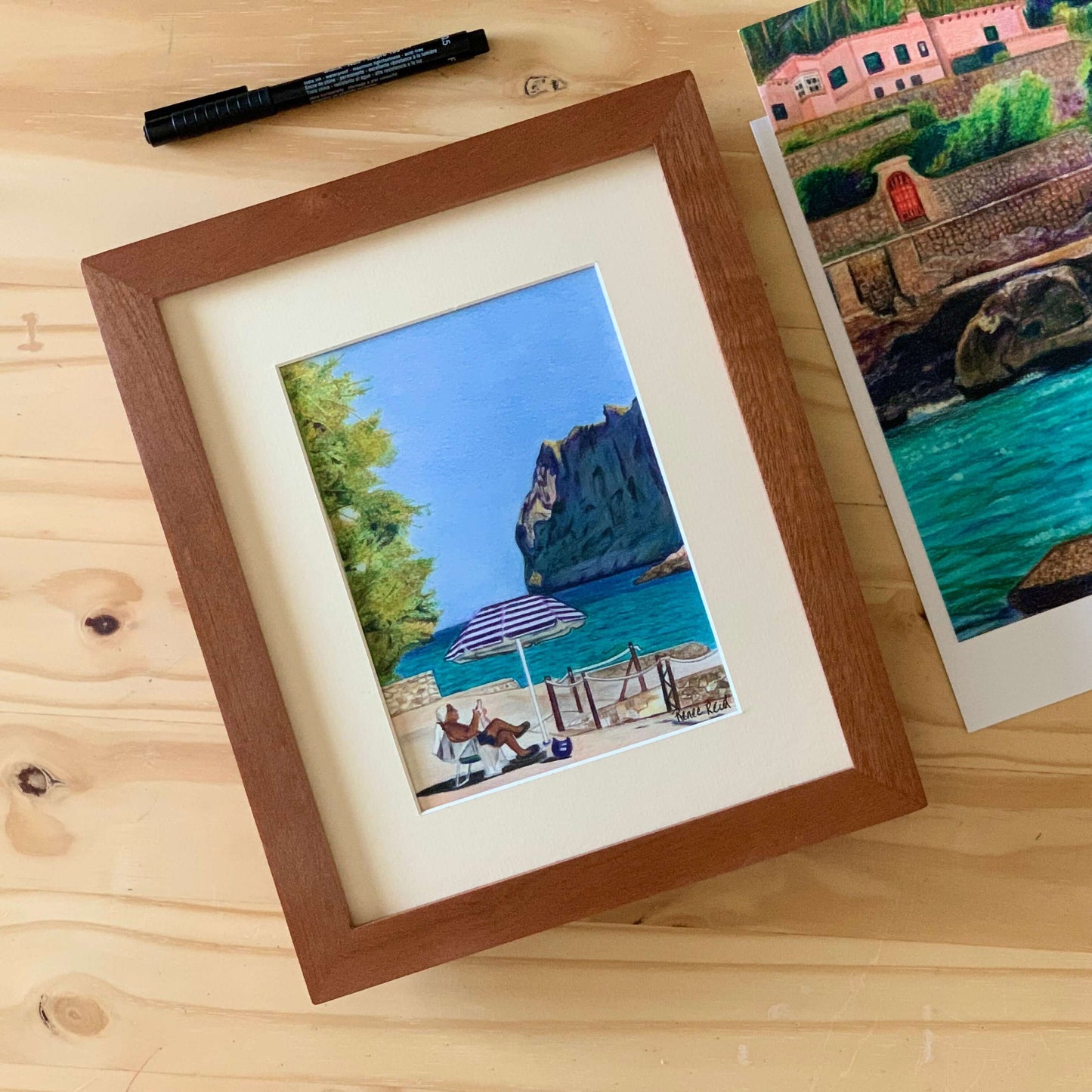 Renee Reid framed original art of a man sitting by the beach under an umbrella reading the newspaper