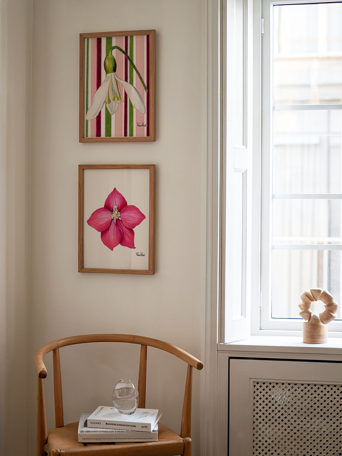 Two framed floral prints on a wall above a wooden chair with books and a glass object.