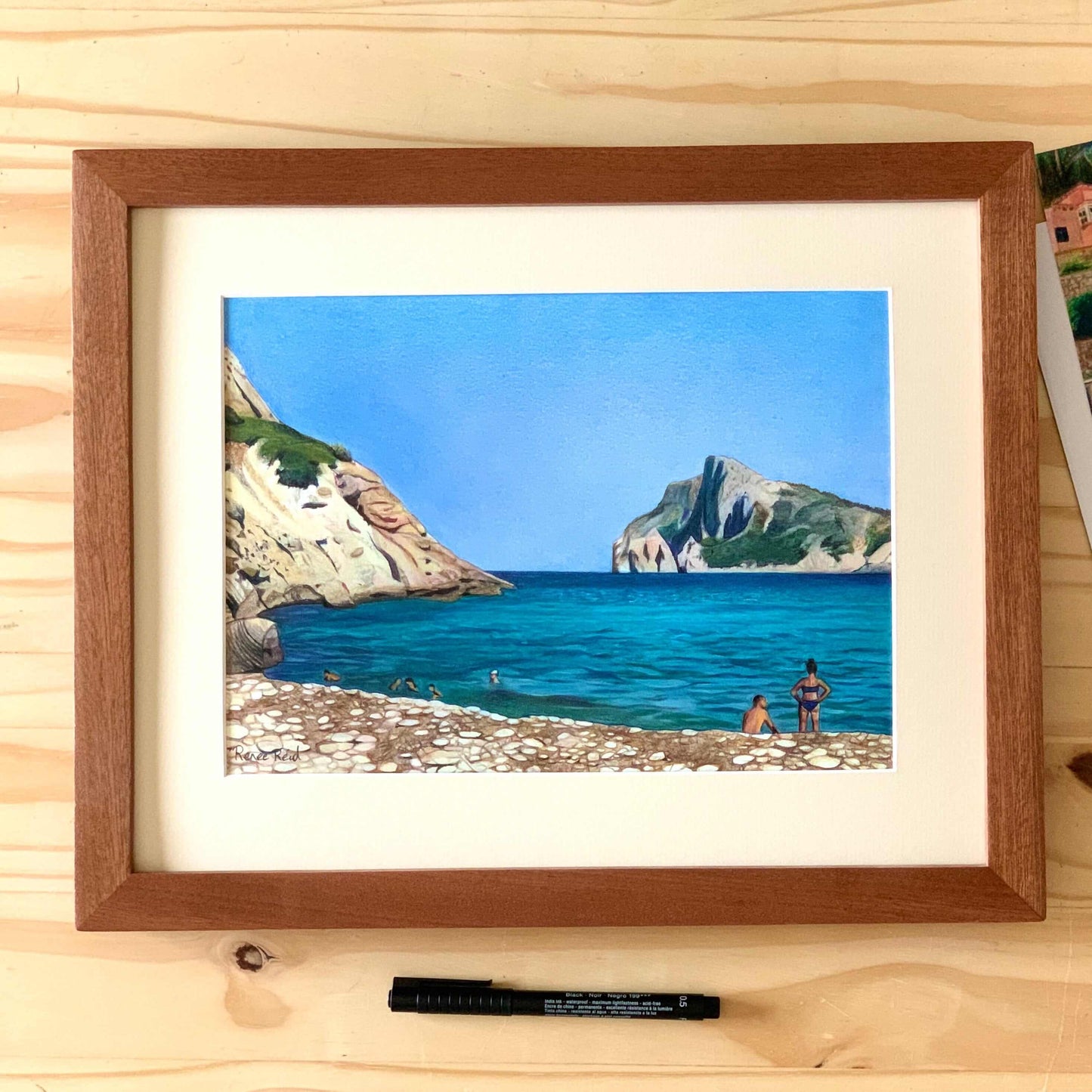 Renee Reid wood framed original art of a beach drawing that has a woman, a man, rocky beach, blue sky and cliffs