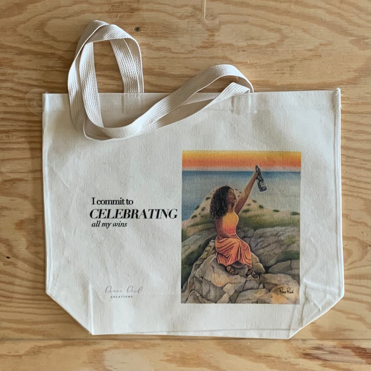 A cream-colored canvas tote bag with white fabric handles, featuring printed text that reads 'I commit to CELEBRATING all my sins' next to an illustration of a woman sitting on rocks by the sea at sunset. The bag is laid flat on a light-colored wooden surface.