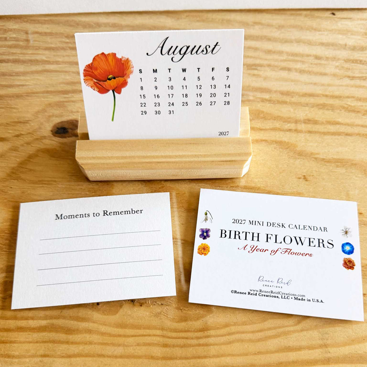 A mini desk calendar set displayed on a wooden tabletop, including a wooden stand, an August calendar page with an red Poppy flower illustration, a 'Moments to Remember' note card, and a '2027 Mini Desk Calendar Birth Flowers' package insert.