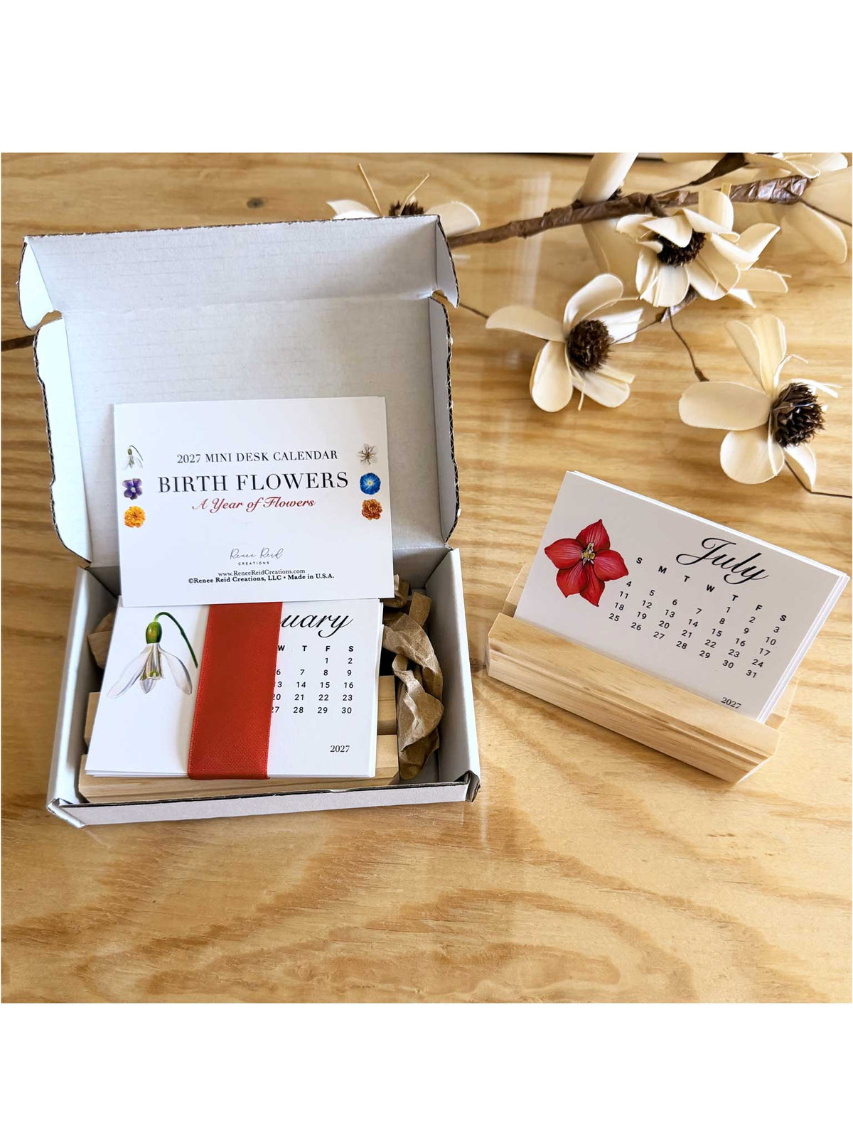 A boxed monthly flower calendar displayed on a wooden surface, with the open box showing a 'Birth Flowers' calendar page and a separate July card resting on a small wooden stand. White flower stems are arranged decoratively in the background.