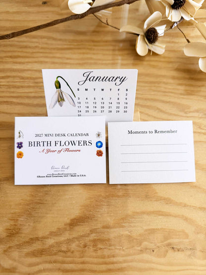 A mini desk calendar set displayed on a wooden tabletop, featuring a January calendar card with a snowflake flower, a front cover card labeled '2027 Mini Desk Calendar Birth Flowers & Year of Flower,' and a 'Moments to Remember' note card. Decorative flowers appear at the top of the frame.