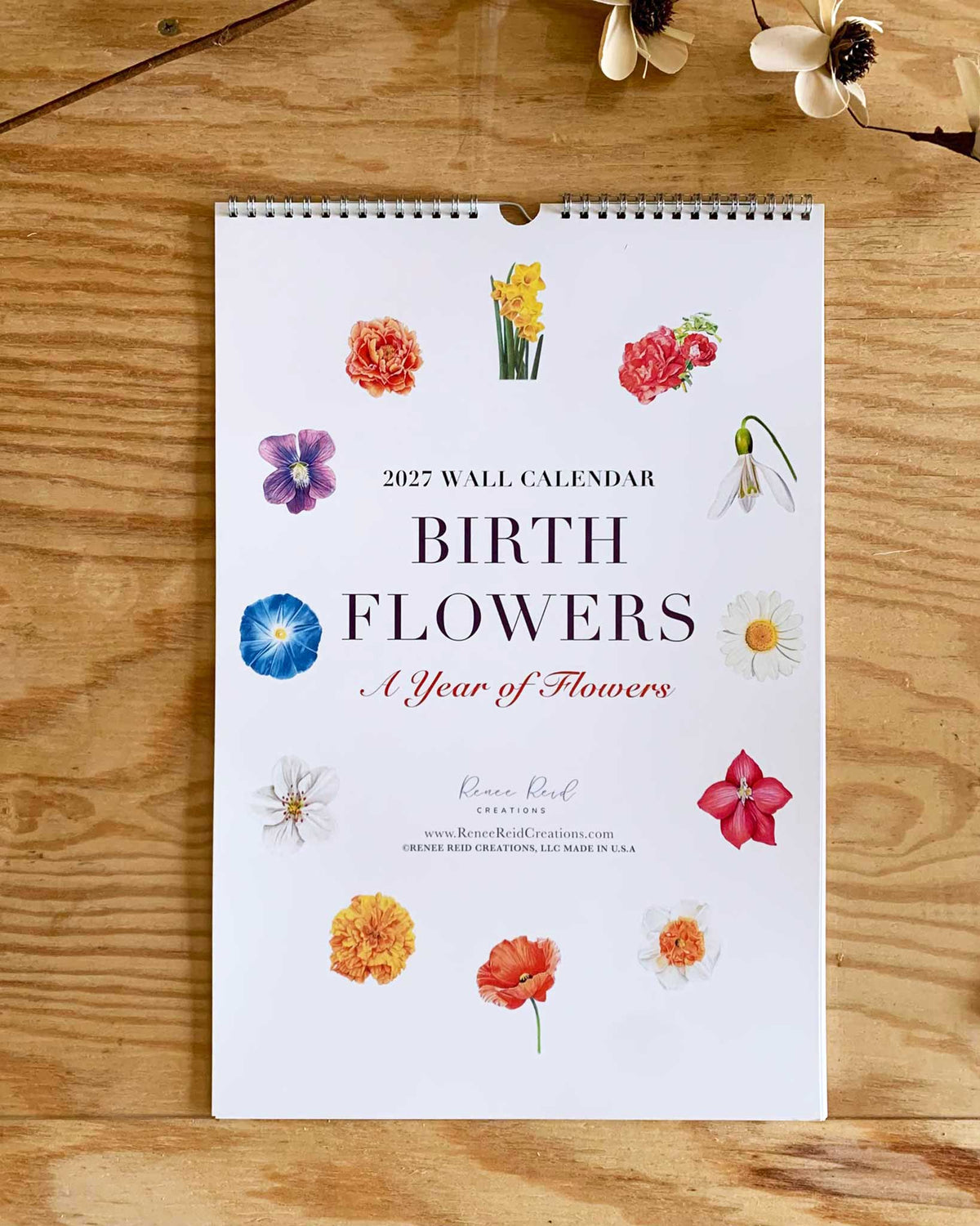A spiral-bound 2027 wall calendar titled 'BIRTH Flowers - A Year of Flowers' by artist Renee Reid  lies flat on a wooden surface. The white cover features colored pencil drawings of various flowers arranged around the title, with small dried flowers placed near the top corners.