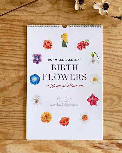 A spiral-bound 2027 wall calendar titled 'BIRTH Flowers - A Year of Flowers' by artist Renee Reid  lies flat on a wooden surface. The white cover features colored pencil drawings of various flowers arranged around the title, with small dried flowers placed near the top corners.