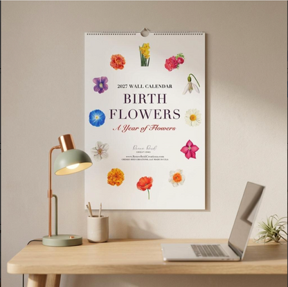 A 2027 wall calendar titled 'BIRTH FLOWERS: A Year of Flowers' hangs on a beige wall above a wooden desk with a green lamp, cup, and laptop. The calendar features a white background decorated with colorful illustrations of various flowers.