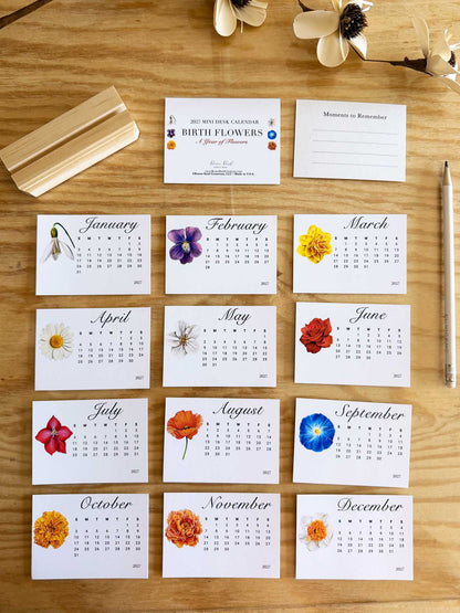 A flat-lay view of a floral monthly calendar on a wooden tabletop, accompanied by a wooden stand, a pen, and dried flowers. The calendar features 12 white cards, one for each month, decorated with different colorful flowers and printed with the month name and dates.
