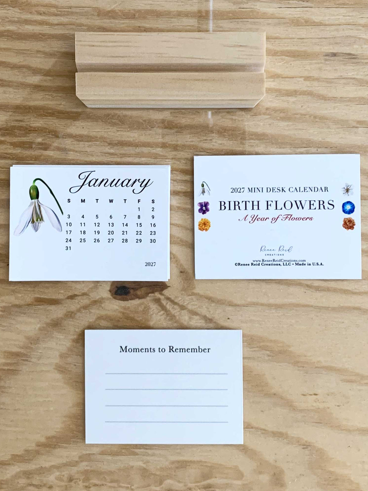 A mini desk calendar set displayed on a light wood background, featuring a January calendar page with a snowball flower illustration, a cover card for a 2027 mini desk calendar titled 'Birth Flowers & Year of Flowers,' and a blank 'Moments to Remember' note card.