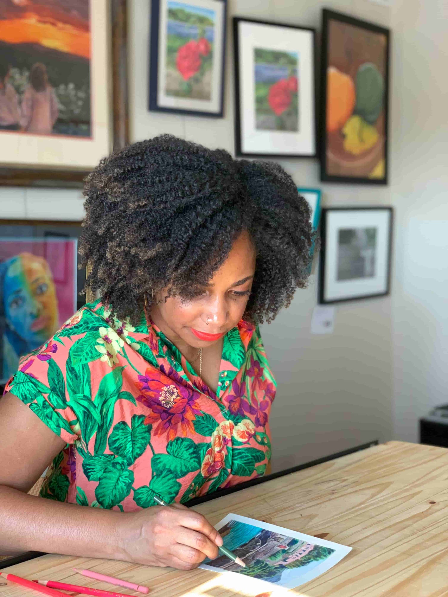 Artist Renee Reid in a colorful shirt painting at a table with framed artwork on the wall.