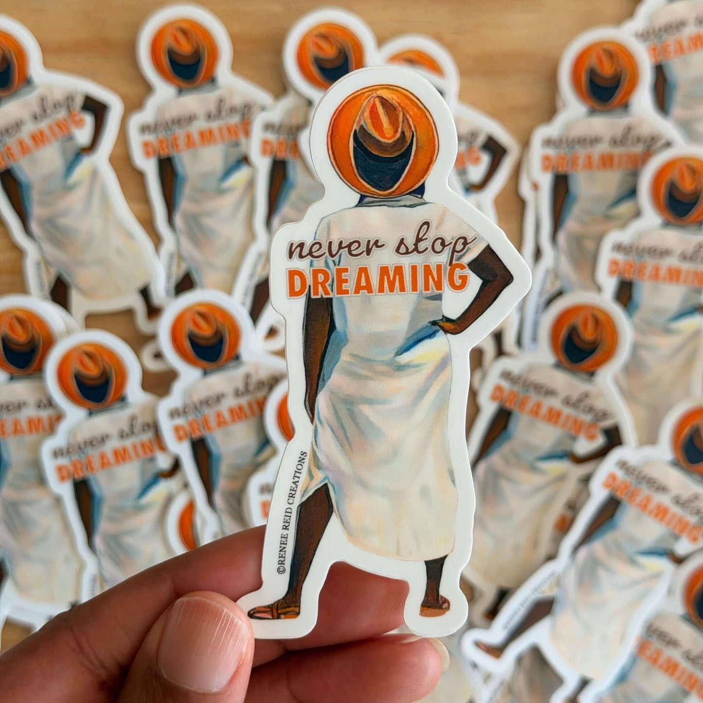 Beyond the Horizon Vinyl Sticker with 'never stop dreaming' text held by a hand, surrounded by more stickers on a wooden surface.