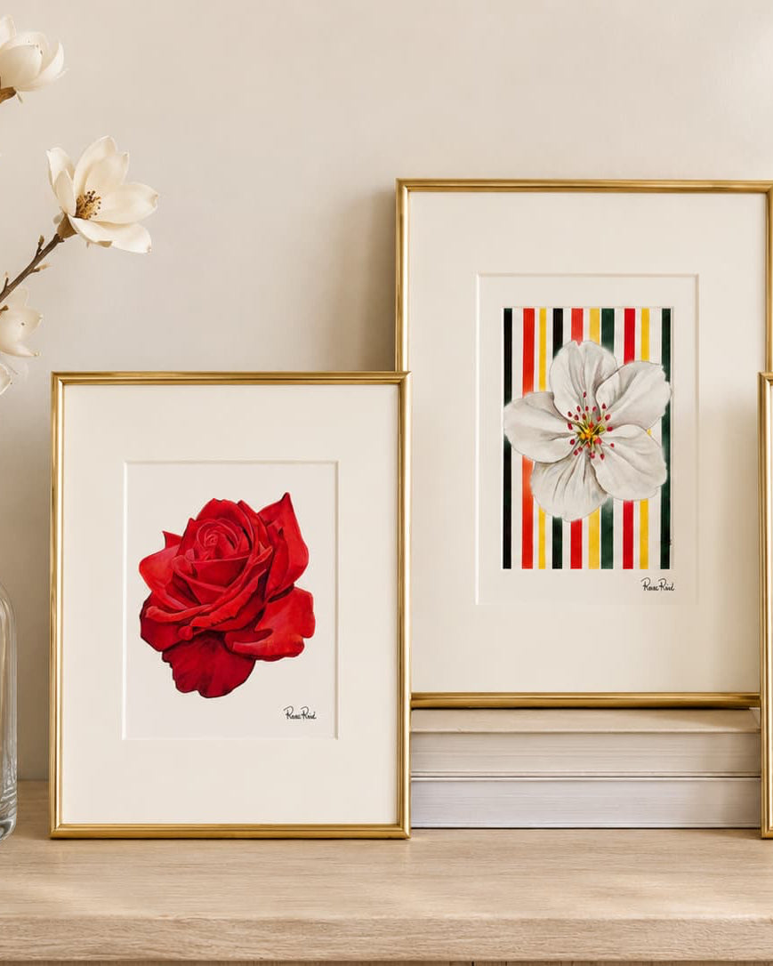 Three framed floral art prints with gold frames are displayed on a light wood surface against a warm beige wall. The center print shows a white flower on vertical rainbow stripes, while the side prints feature a red rose and an orange marigold.