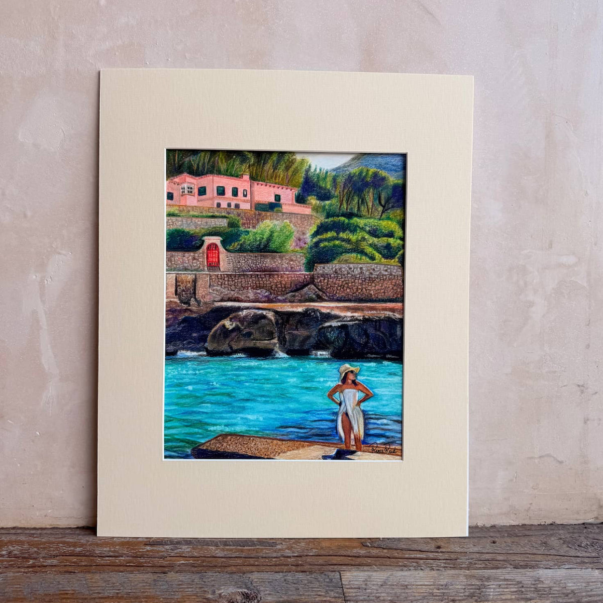 A matted art print from Renee Reid Creations featuring a colorful landscape painting of a coastal scene with turquoise water, rocks, greenery, and pink buildings, displayed against a textured beige wall on a rustic wooden surface.