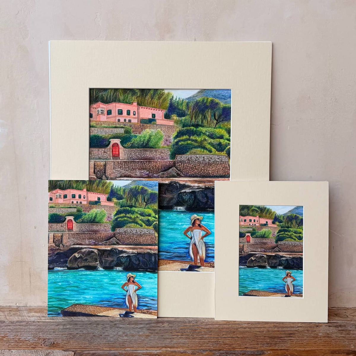 Three matted art prints from Renee Reid Creations featuring a colorful landscape painting of a coastal village with turquoise water, displayed on a rustic wooden surface against a textured beige wall.