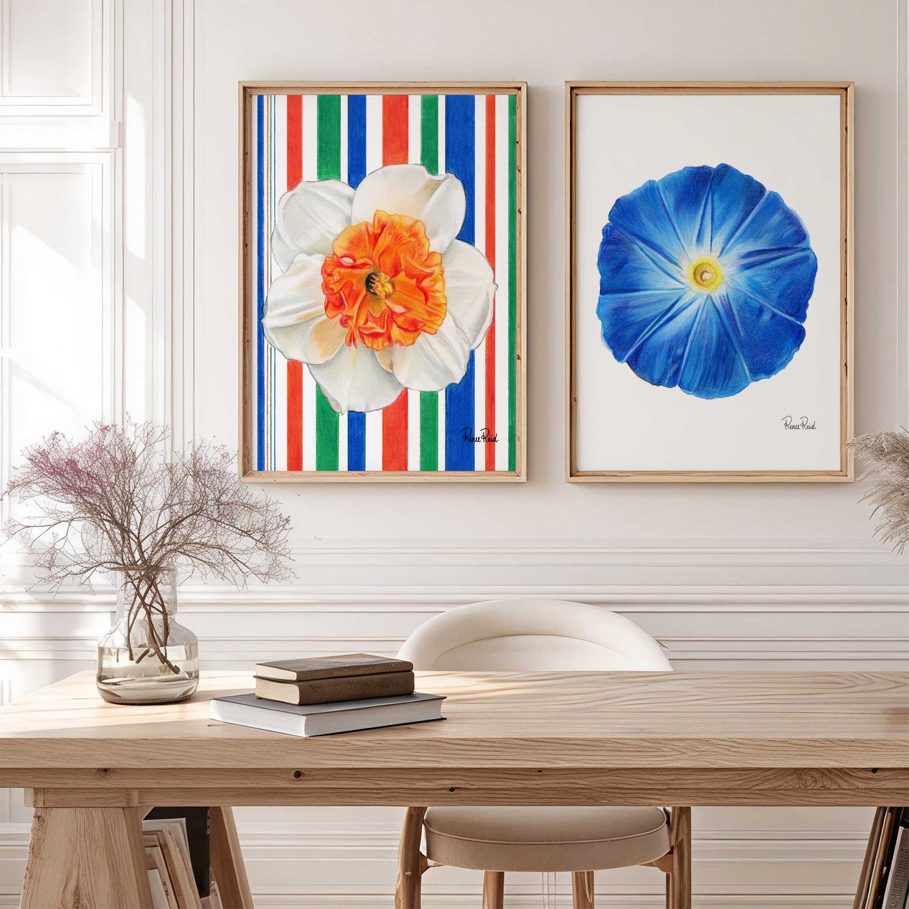 Two floral artworks on a wall above a wooden table with books and a vase.