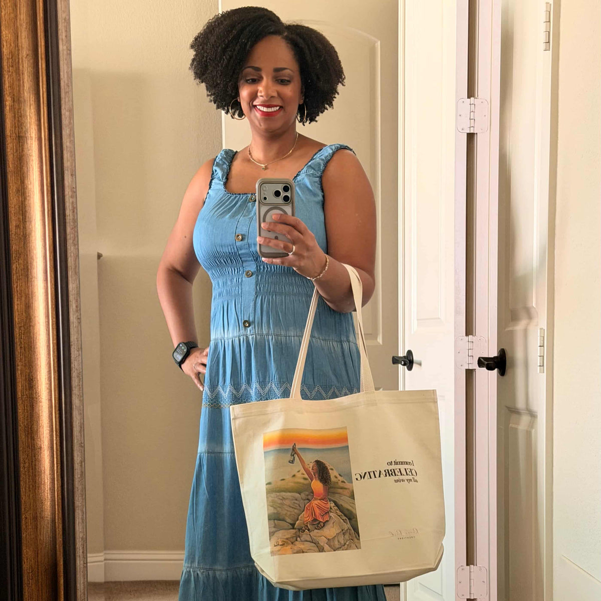 A woman takes a mirror selfie in a home hallway, wearing a light blue tiered midi dress and holding a cream canvas tote bag with a colorful graphic print that says I commit to CELEBRATING all my wins. She is wearing yellow slip-on shoes.