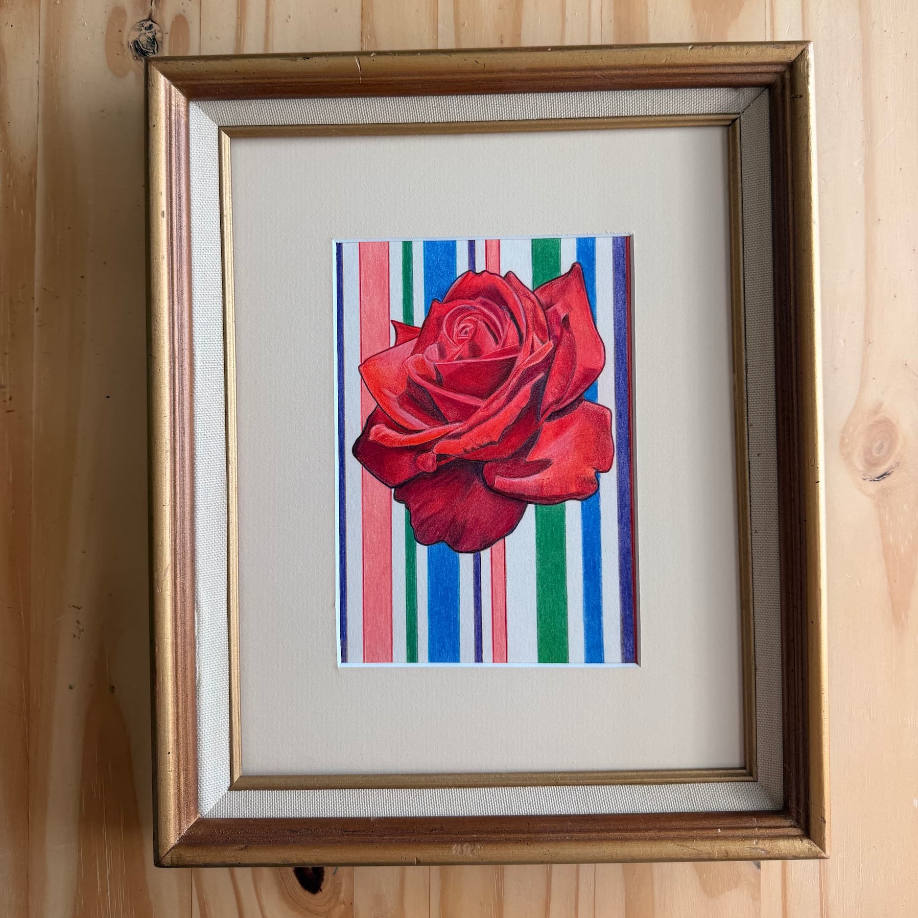 Framed artwork of a red rose on a striped background against a wooden wall.