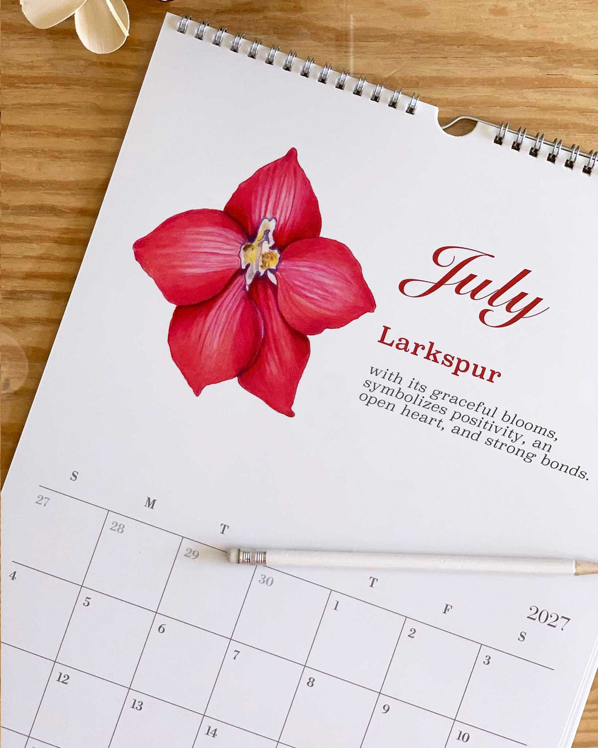 A spiral-bound July calendar page with a large red flower illustration and the title 'July Larkspur,' placed on a wooden surface with a white pen resting across it.