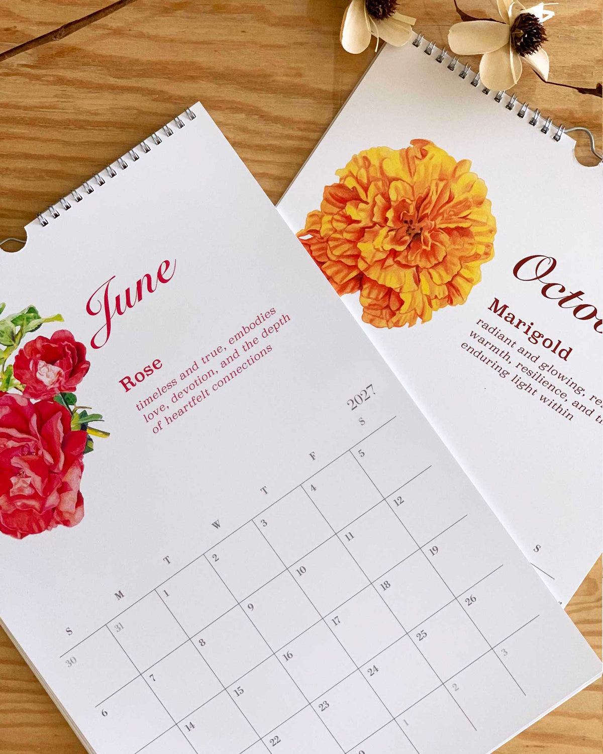 Two spiral-bound monthly calendar pages laid on a wooden surface, featuring floral illustrations and month names June and October. The pages are white with red and orange floral artwork and decorative flowers at the top.