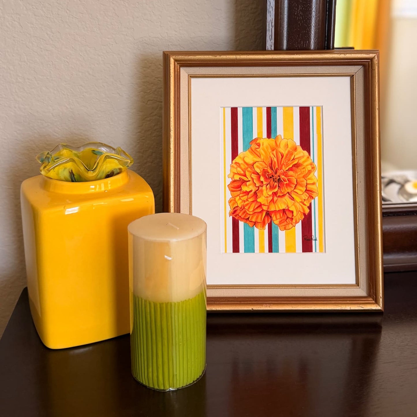 A framed 5x7 art print featuring a vibrant orange marigold flower on a background of colorful vertical stripes, styled on a dark wooden table next to a yellow container and a green candle.