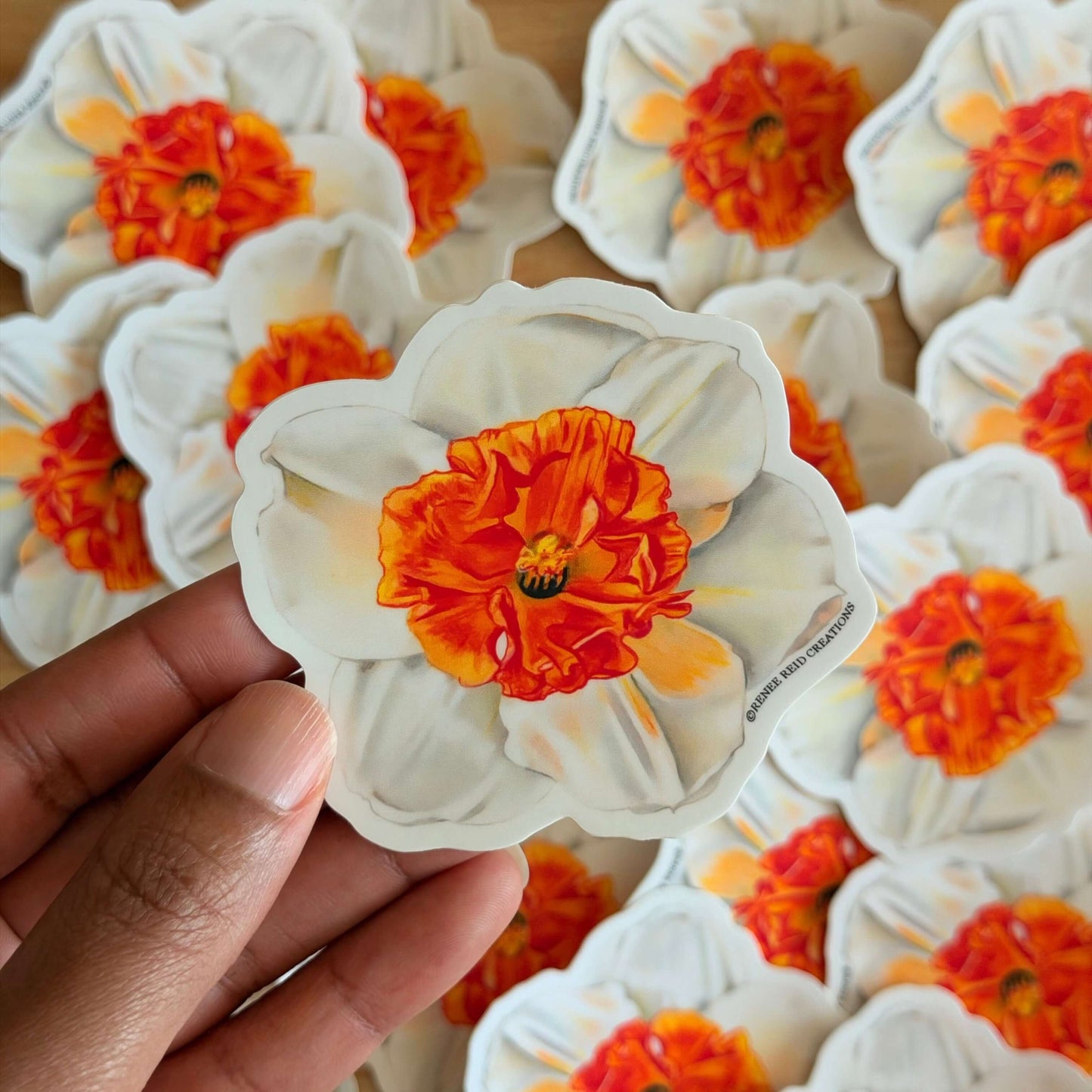 Hand holding a fOrange Narcissus Flower Vinyl Sticker, surrounded by more stickers on a wooden surface.