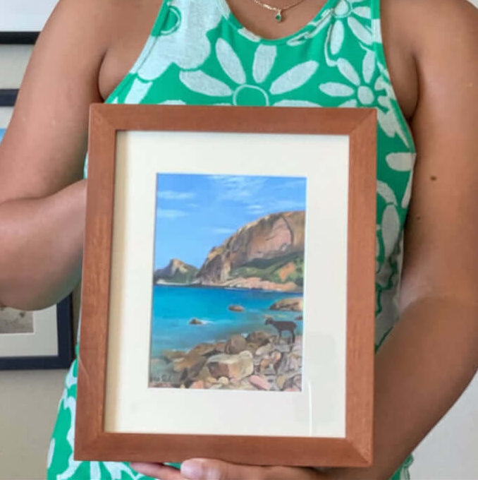 Artist Renee Reid holding a framed artwork of a coastal scene in an art gallery.