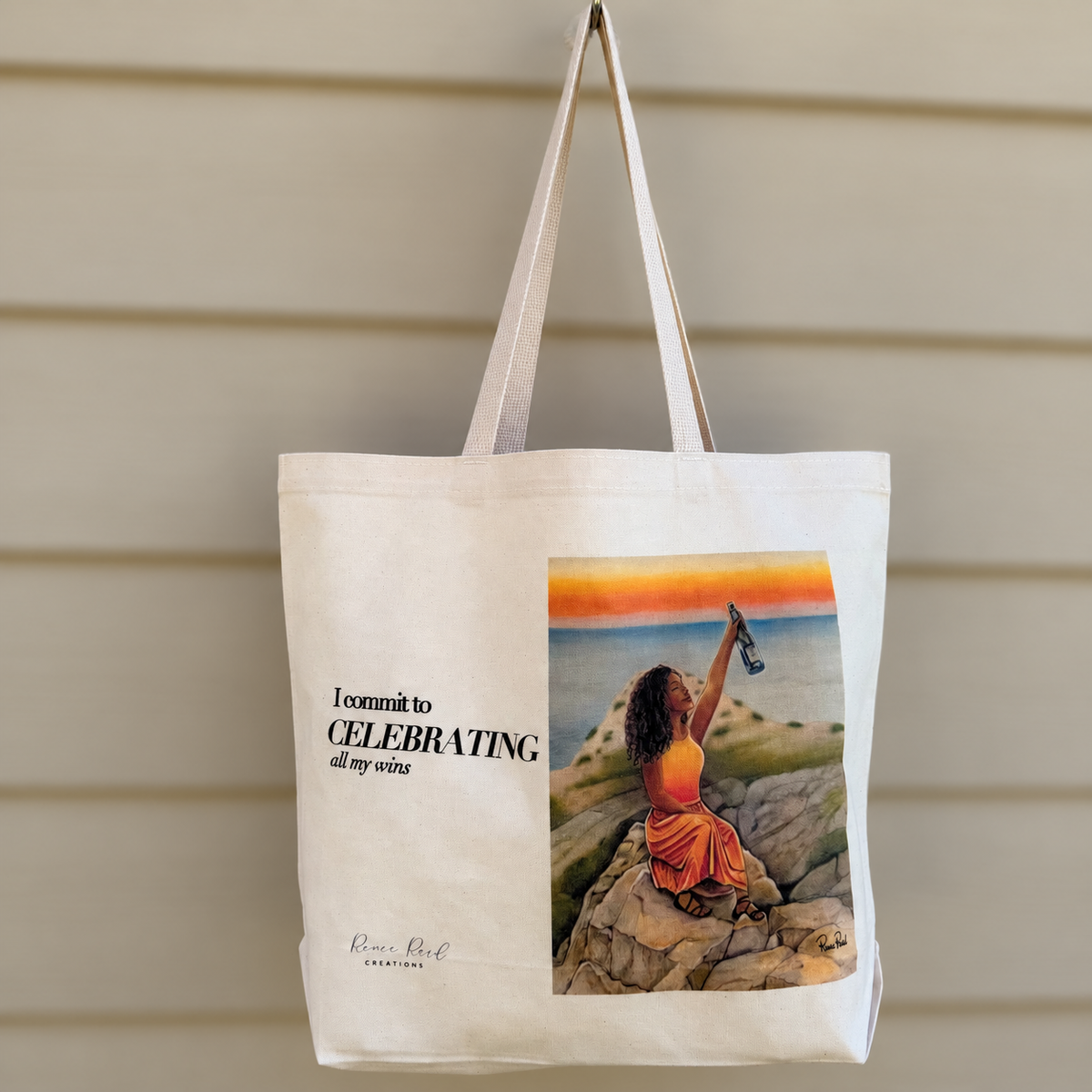 A cream canvas tote bag with dual shoulder straps, hanging against a beige wood-paneled wall. The bag features a printed graphic of a woman in an orange dress sitting on rocks by the sea at sunset, alongside the text 'I commit to CELEBRATING all my sins' and the brand name 'Pura Lip Design'.