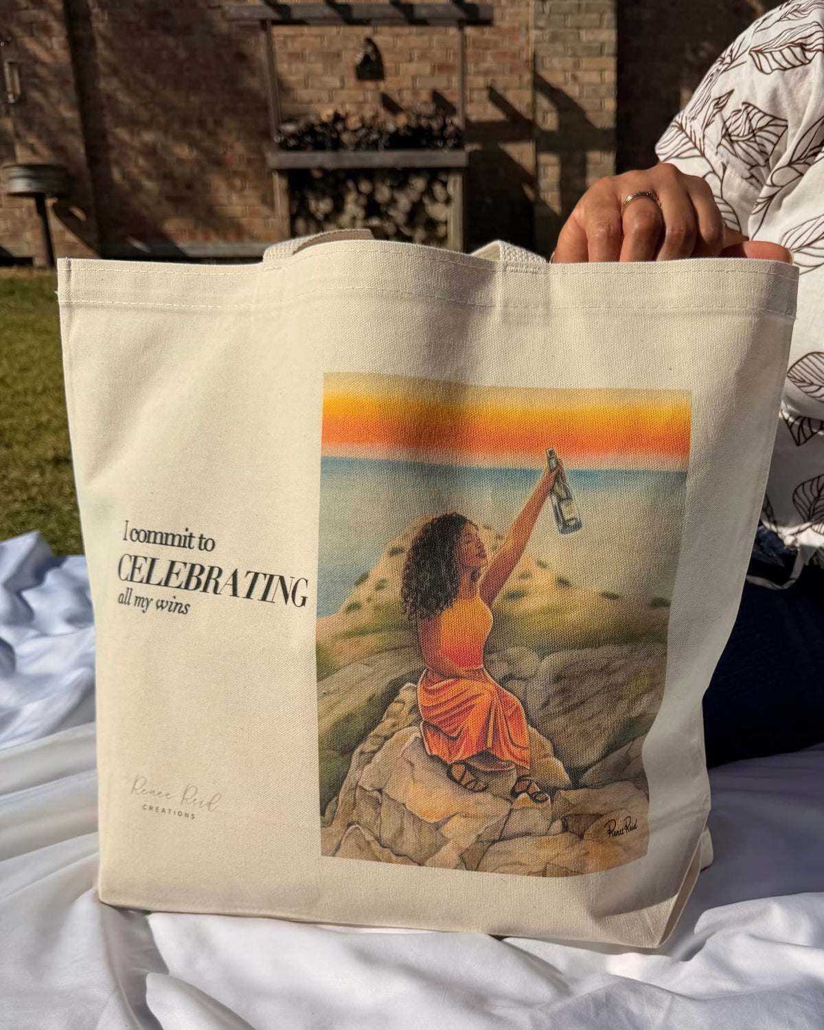 A cream canvas tote bag with printed text and an illustration of a woman on a rock by the sea with “I commit to CELEBRATING all my wins”, held by a person outdoors on a white blanket.