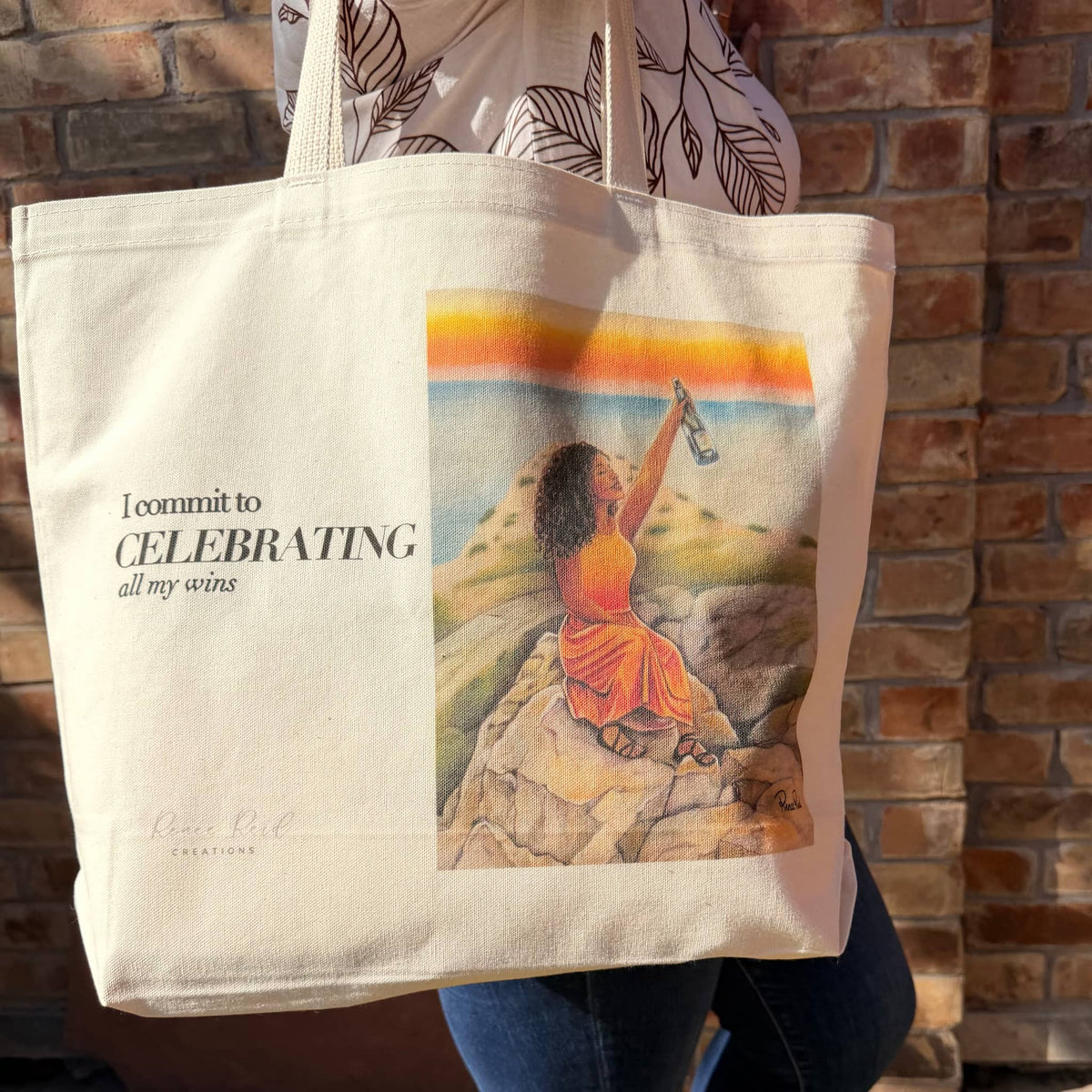 A woman holds a large cream canvas tote bag with printed text reading 'I commit to CELEBRATING all my wins' and a colorful illustration of a seated woman on rocks by a beach. The tote bag is shown against a brick wall background.