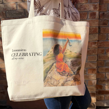 A woman holds a large cream canvas tote bag with printed text reading 'I commit to CELEBRATING all my wins' and a colorful illustration of a seated woman on rocks by a beach. The tote bag is shown against a brick wall background.