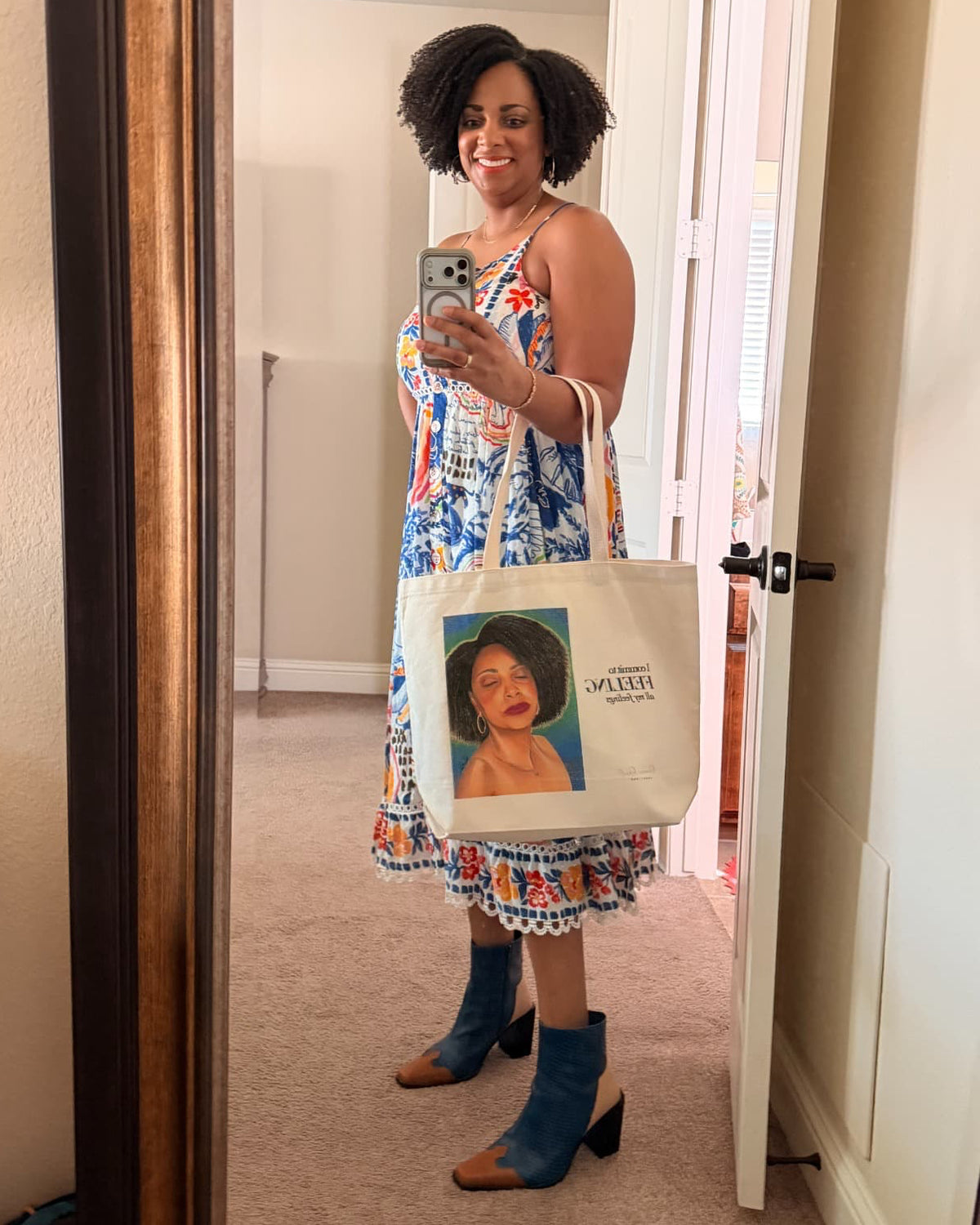 A woman takes a mirror selfie in a hallway, wearing a colorful floral print dress and blue heeled boots, while holding a canvas tote bag with a printed portrait and the words 'I commit to FEELING all my feelings'.