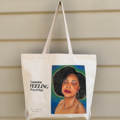 A cream canvas tote bag hanging against beige siding, featuring a printed portrait of a woman with dark hair and the text 'I commit to FEELING all my feelings'.