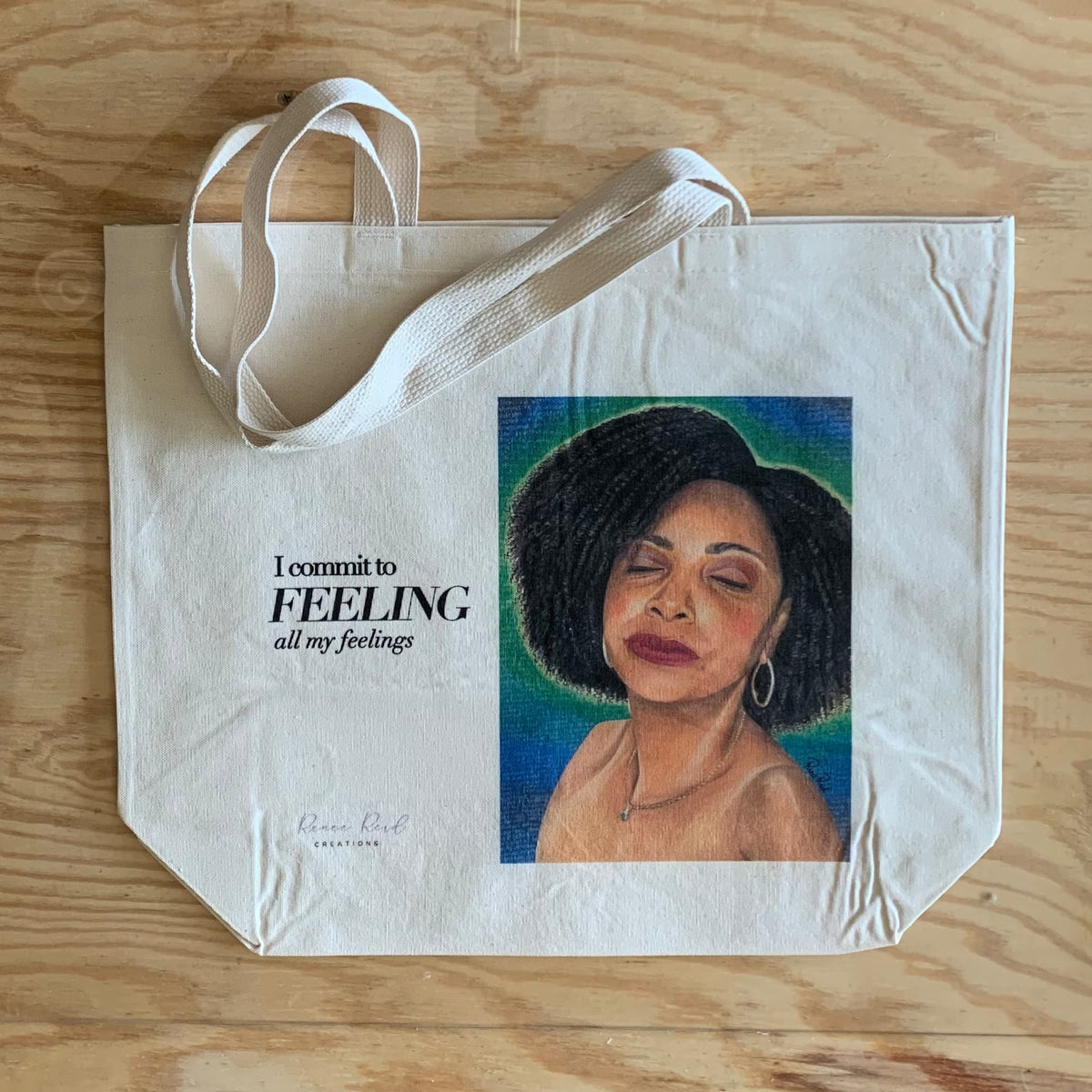A cream-colored canvas tote bag with white handles, featuring a printed portrait of a woman with black hair against a blue and green background, next to the text 'I commit to FEELING all my feelings'. The bag is laid flat on a light wood grain surface.