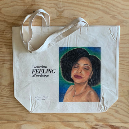 A cream-colored canvas tote bag with white handles, featuring a printed portrait of a woman with black hair against a blue and green background, next to the text 'I commit to FEELING all my feelings'. The bag is laid flat on a light wood grain surface.