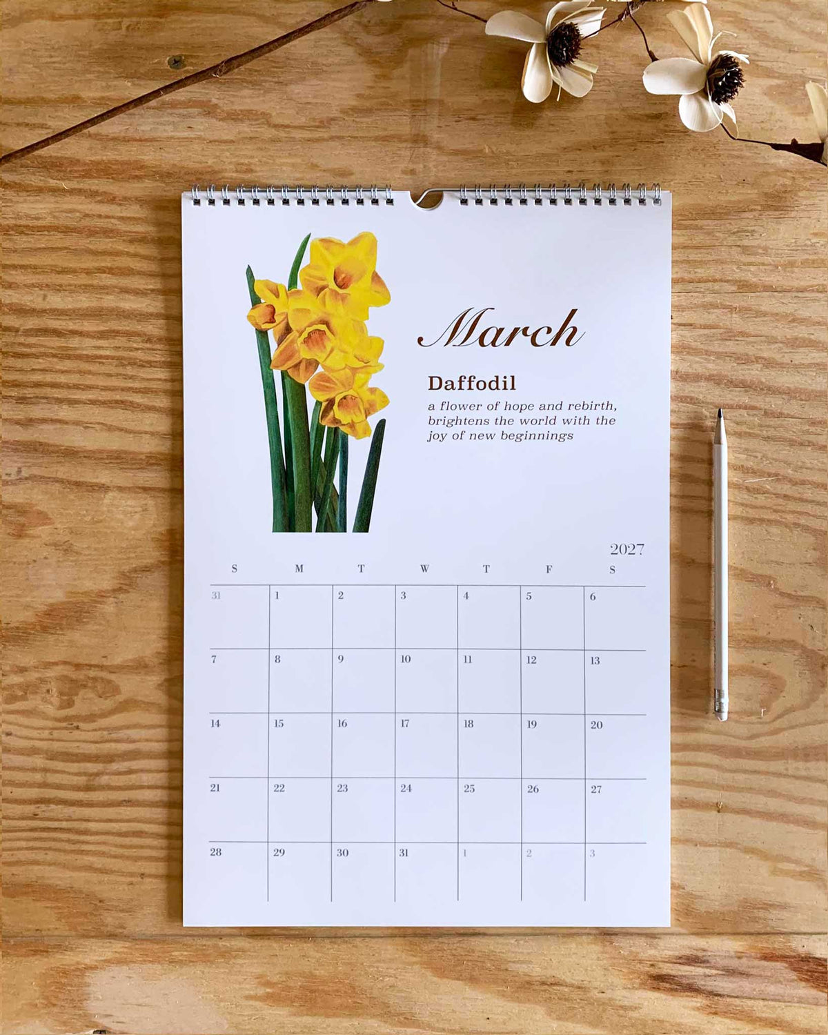 A March daffodil calendar page laid flat on a wooden surface, with a photo of yellow daffodils, the month name in script, a short description, and a 2017 weekly calendar grid. A pencil and small flowers are placed nearby as props.