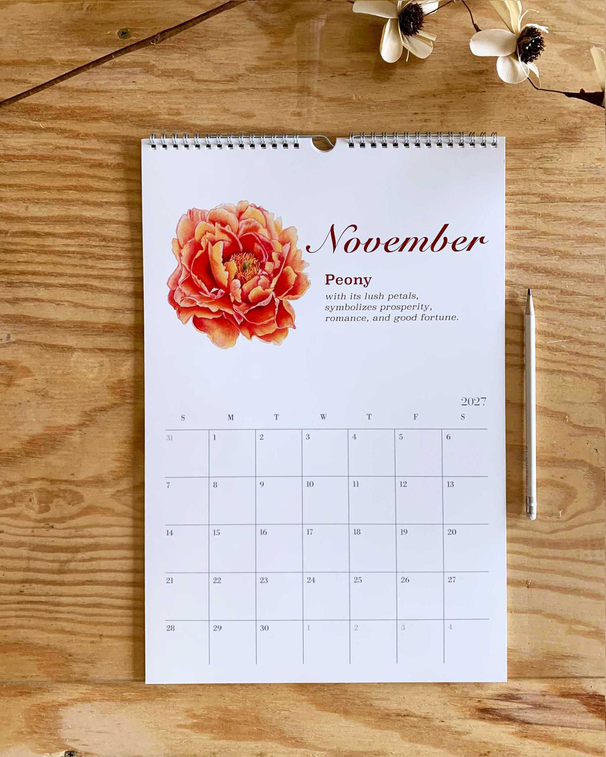 A spiral-bound November desk calendar page featuring a large peach and orange peony flower with the word 'November' and a short poem, placed on a wooden tabletop with a pencil and small flowers nearby.
